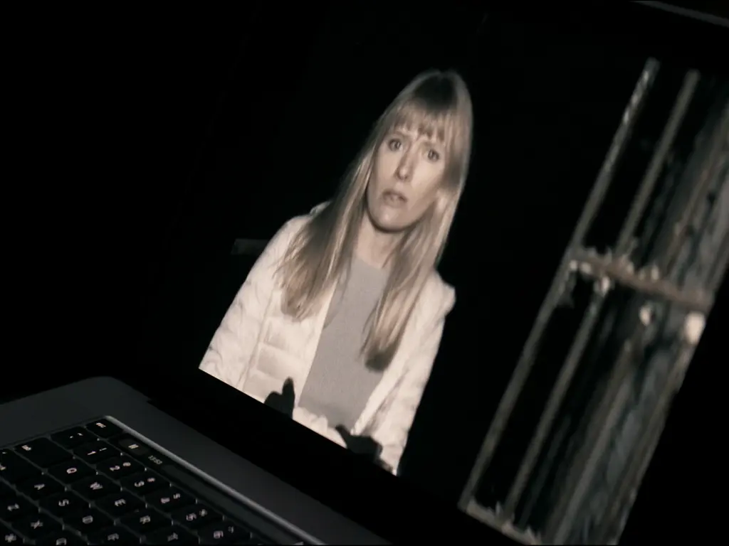 Blonde woman with fringe displayed on laptop screen, second laptop visible to the right, dark background.