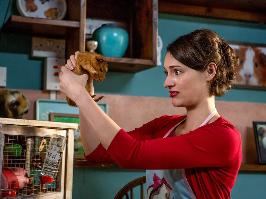 A woman in a red dress holds up a small furry animal in a cluttered room with shelves and ornaments.
